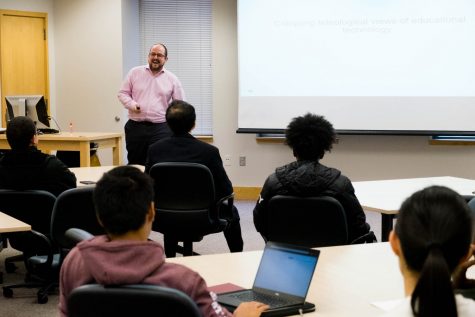 A man in a classroom laughs as he looks at those seated at tables.