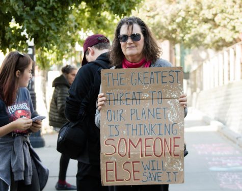 A woman holds a cardboard sign reading "The greatest threat to our planet is thinking someone else will save it."