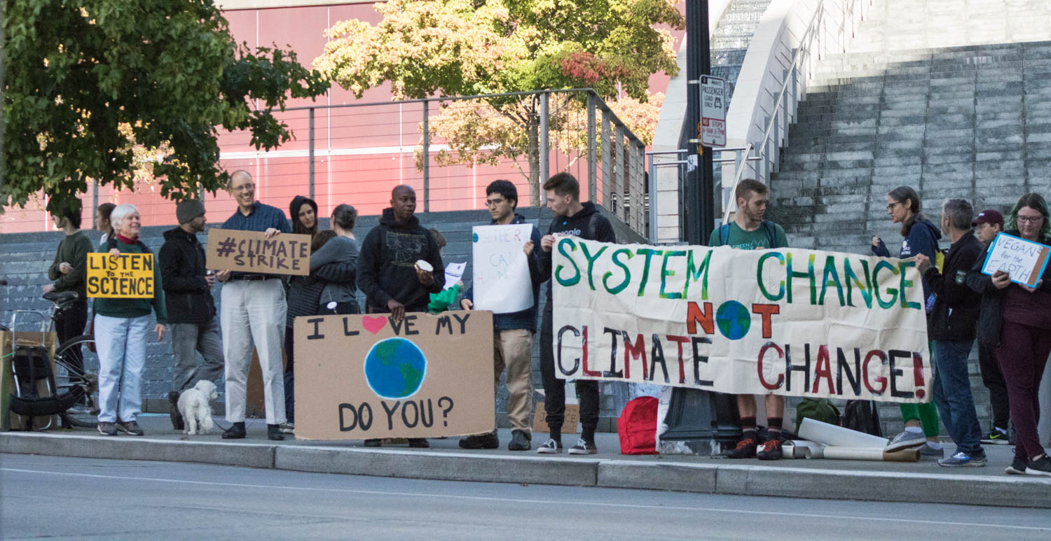 Protesters hold signs along a sidewalk. The signs read "Listen to the science", "#ClimateStrike" , "I love my planet do you?" and "System change not climate change!"
