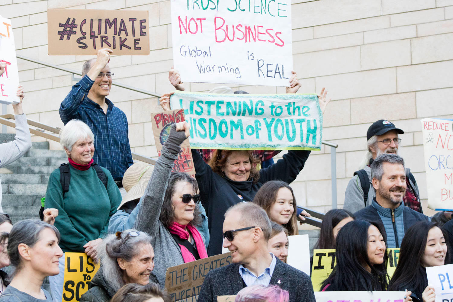 Protesters pose for a group photo on the steps of a building. The signs read "#ClimateStrike", "Trust science not business. Global warming is real" and "Listening to the wisdom of youth."