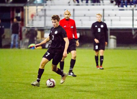 A man in a black Seattle Pacific soccer uniform dribbles a soccer ball toward the foreground. There is a referee in a red uniform and a man in a black SPU uniform behind him.
