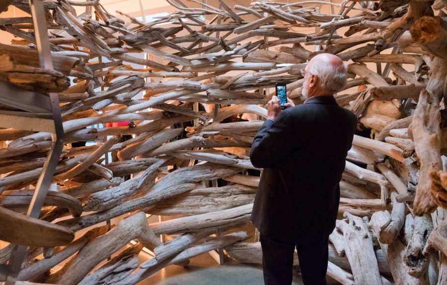 A elderly man takes a picture on his phone while inside a scuplture made of wooden branches.