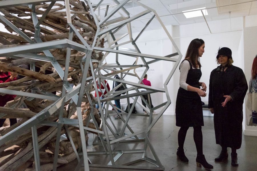 A woman speaks to another woman beside a sculpture made of wood branches and framed in metal.