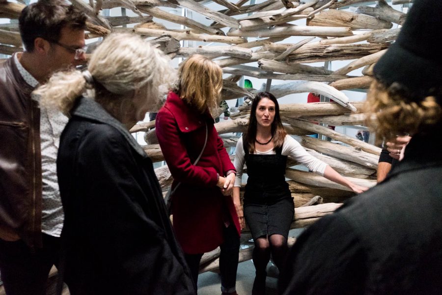 A woman speaks to a group of people within a scupture made of wooden branches.