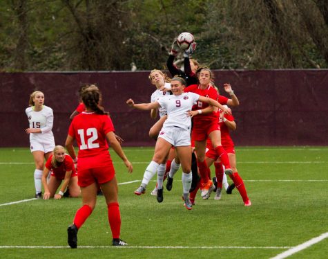 A woman in a white Seattle Pacific soccer uniform tries to head butt a soccer ball as it is grabbed by a woman in a black Simon Fraser goalie uniform. They are surrounded by soccer players in red and white uniforms.