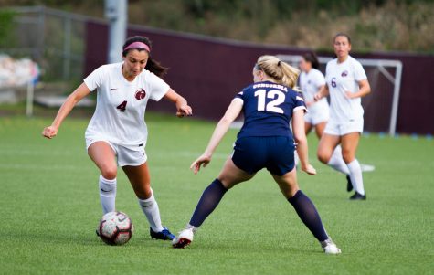 A woman in a white jersey kicks a soccer ball while a woman in a blue jersey lunges to steal the ball