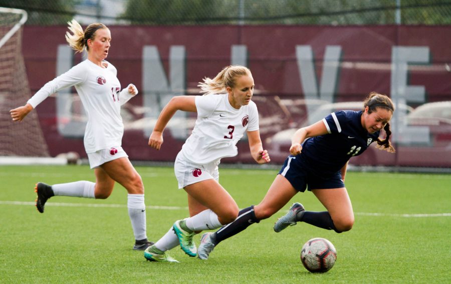 A woman runs forward kicking a soccer ball while another falls to the ground