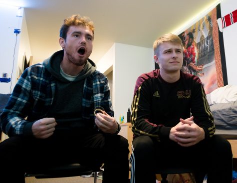 Two men make expressions at a t.v. screen while a sports game plays