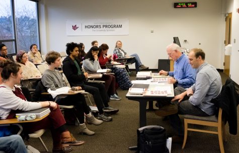 two professors sit at a table in front of a classroom full of student