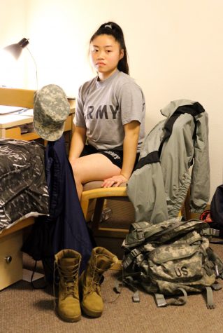 A woman in an "Army" shir sits at a desk with combat boots and a camouflage backpack at her feet.