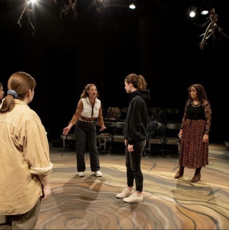 A photo of some of the cast of Everybody (L to R Clara Christensen, Ellie Duenow, Emily Haan, Lia Harper) in their costumes on stage of Everybody. They are all circling around Emily as Emily stands in the center of the stage. 