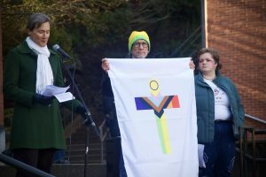Rev. Dr. Koenig speaks while Kevin Neuhouser holds a Christian LGBTQIA+ pride flag, alongside student Gail McMillan.